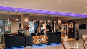 Un espace d’accueil d’hôtel au design moderne, comprenant un comptoir d’accueil, un coin salon et un grand vase rempli de fleurs.