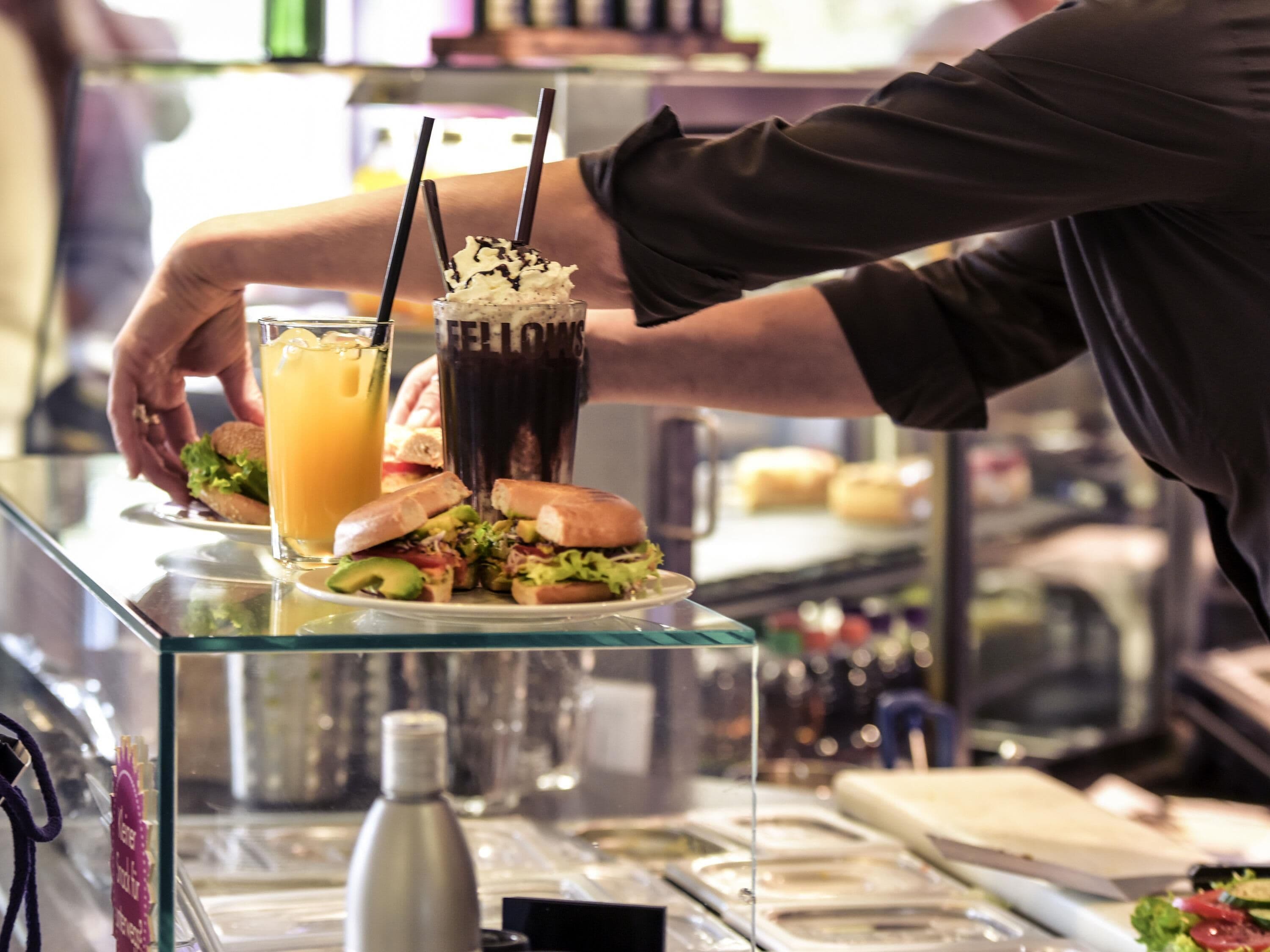 A person serving a sandwich with a drink on a glass counter.