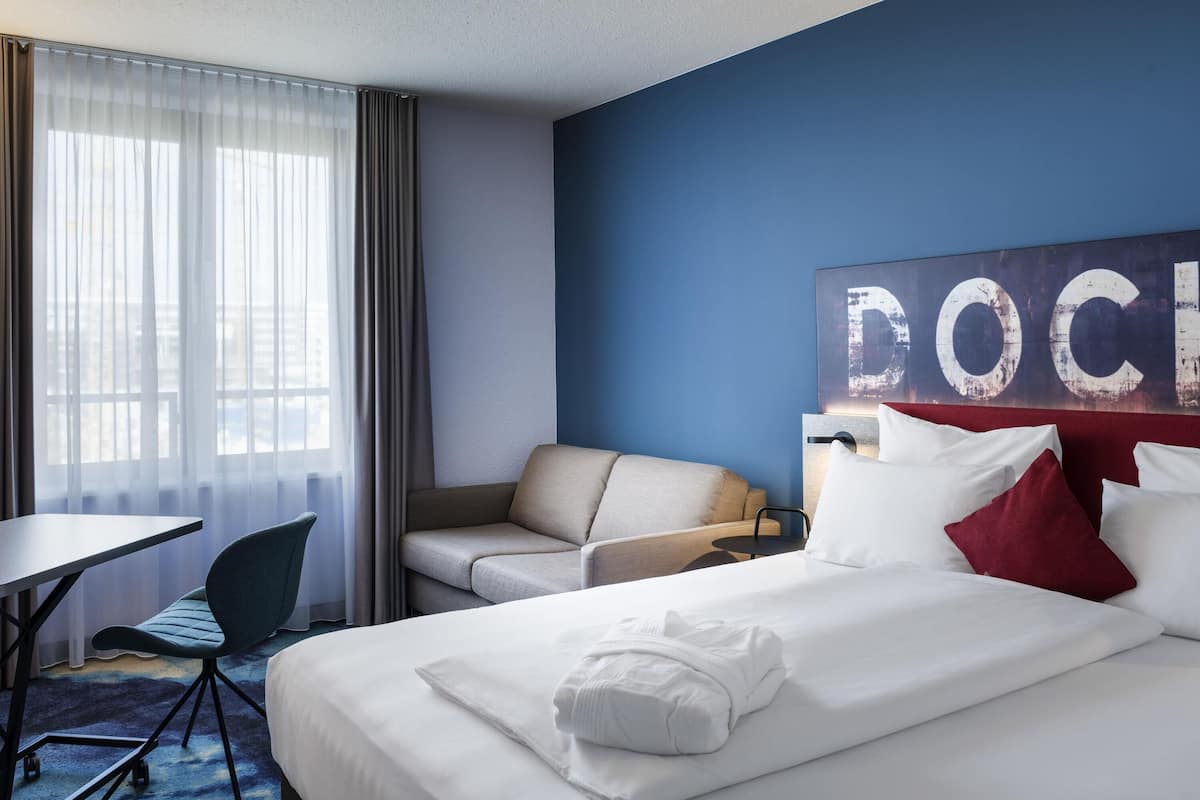 A hotel room with a bed, a desk, a chair, a sofa, and a large wall art piece.