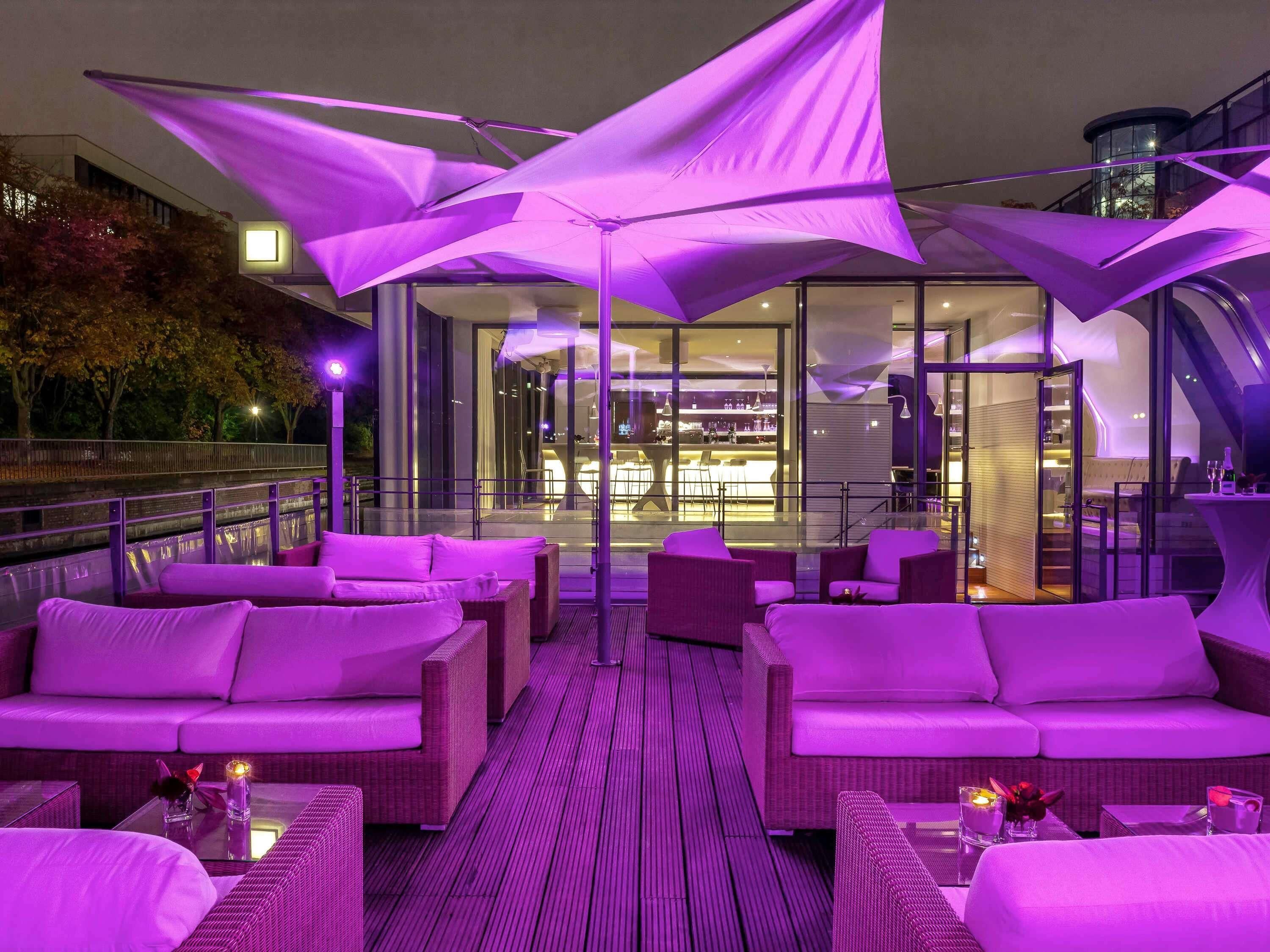 A rooftop bar with purple lighting, wicker furniture, and a view of the cityscape.