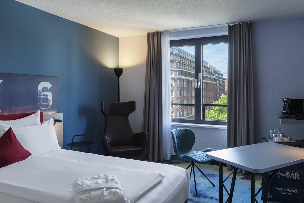 Ett hotellrum med en säng, två stolar, ett bord, en lampa, ett fönster med utsikt över byggnader och en bardisk.