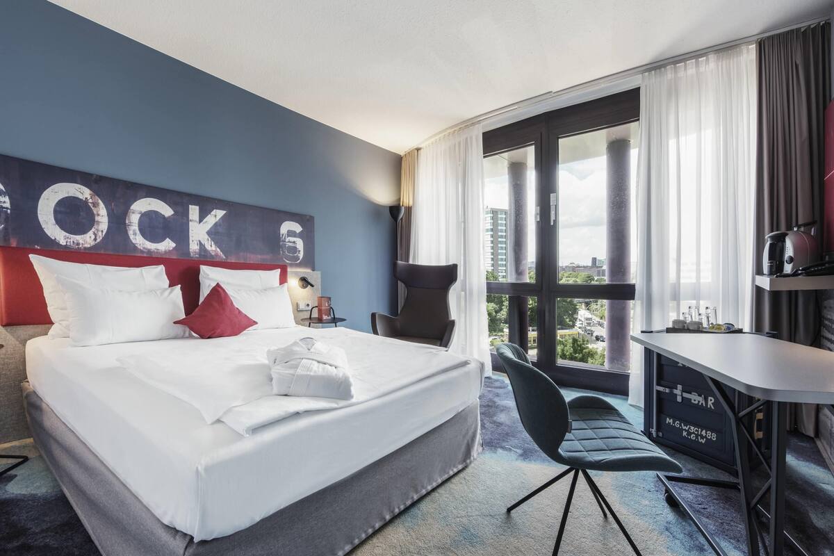 Ein Hotelzimmer mit einem großen Bett, einem Schreibtisch und einem Stuhl. Durch das Fenster ist die Stadt zu sehen.