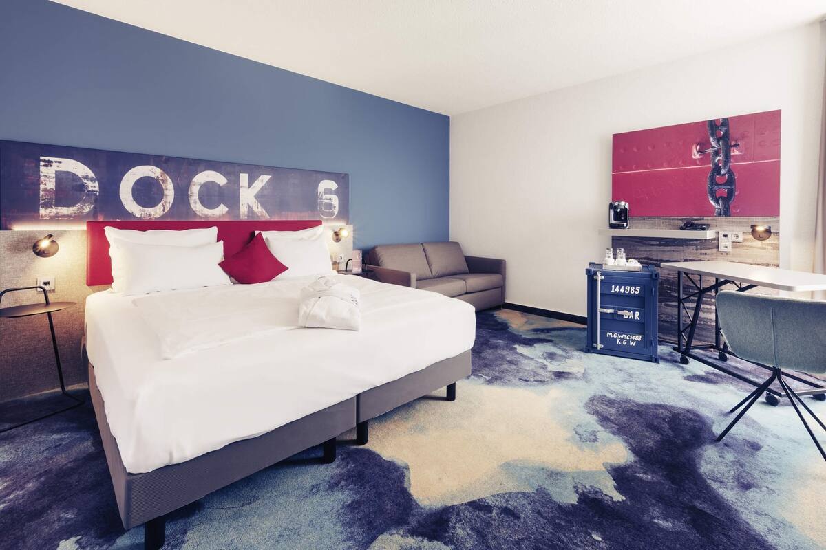 Ein Hotelzimmer mit einem Bett, einem Schreibtisch, einem Sessel, einer Couch, einem wandmontierten Fernseher und einem großen Kunstwerk.