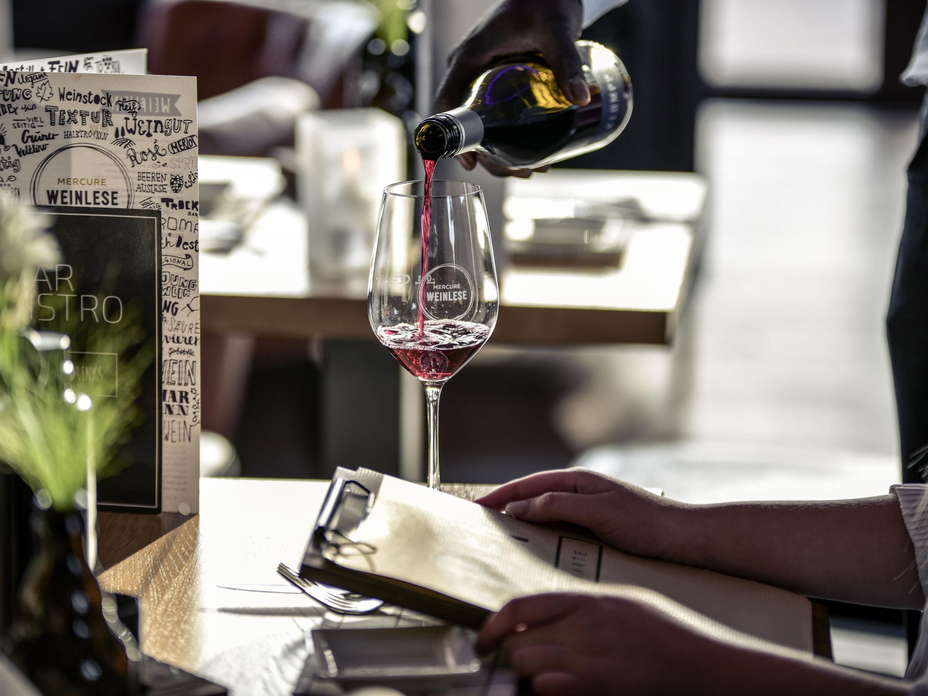 A person pouring wine into a glass, with a menu and a bottle visible on the table.