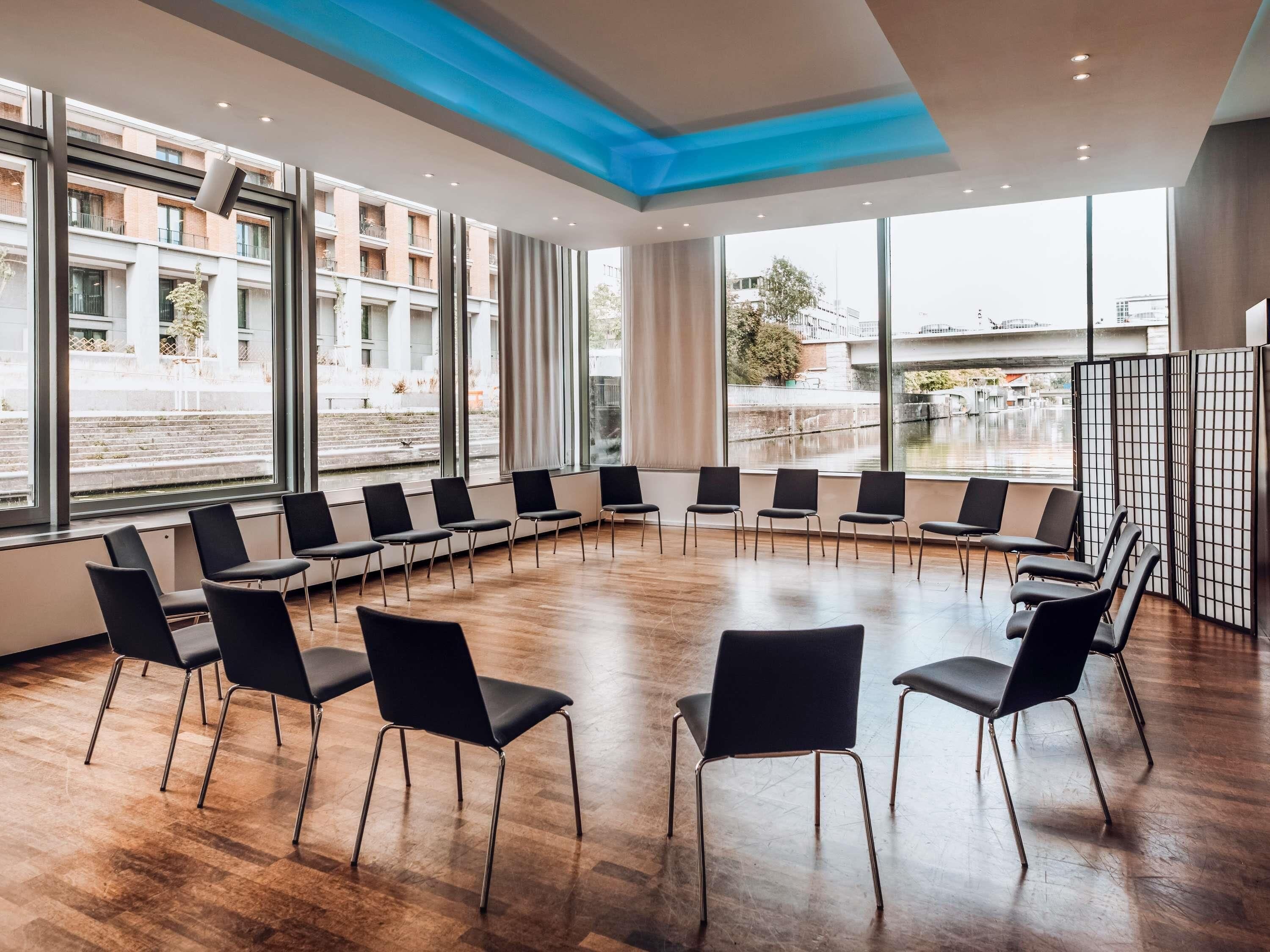A conference room with a circular seating arrangement, large windows, and a view of a building and a canal.