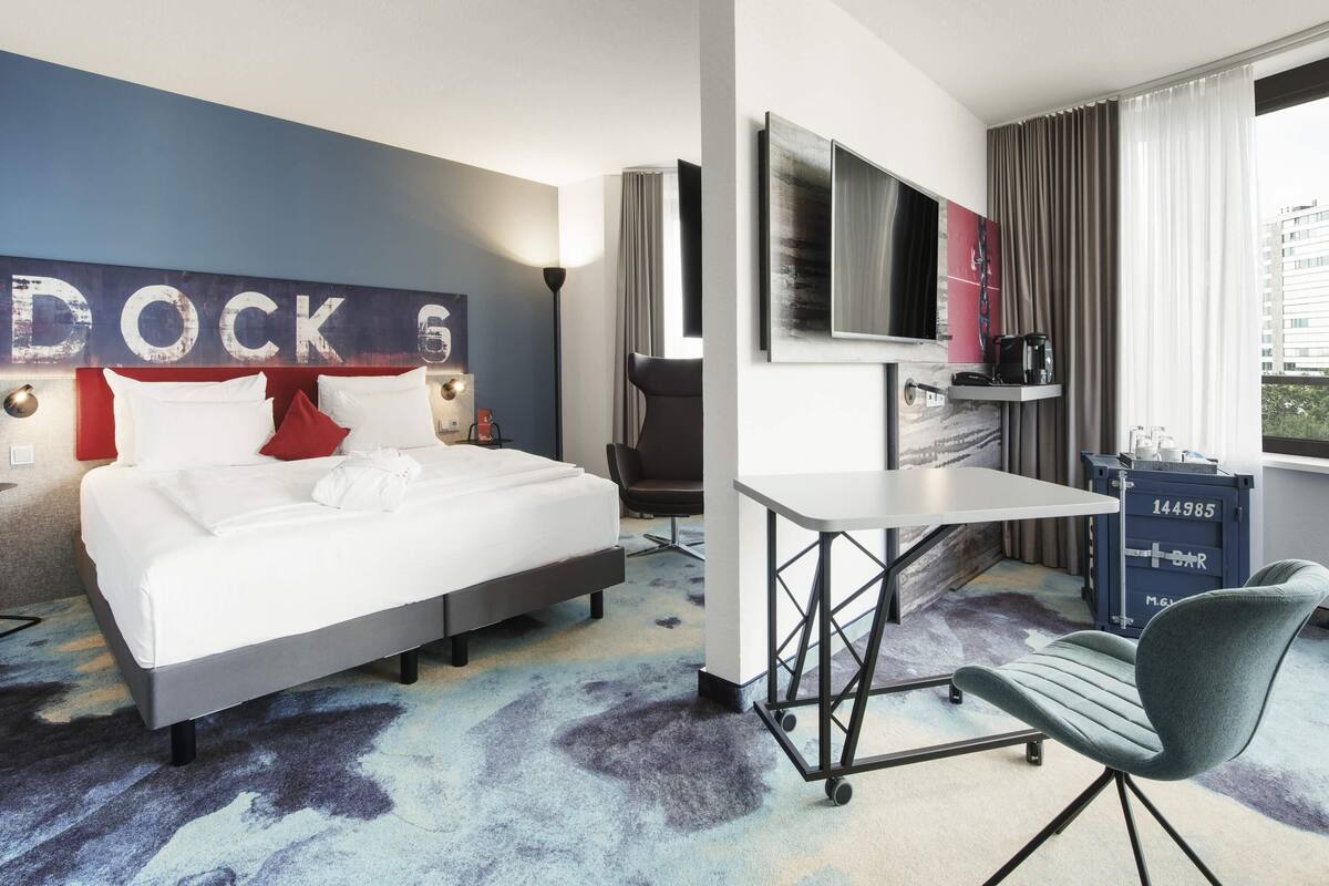 Ein Hotelzimmer mit Bett, Schreibtisch, Stuhl, Fernseher und einem großen Fenster.