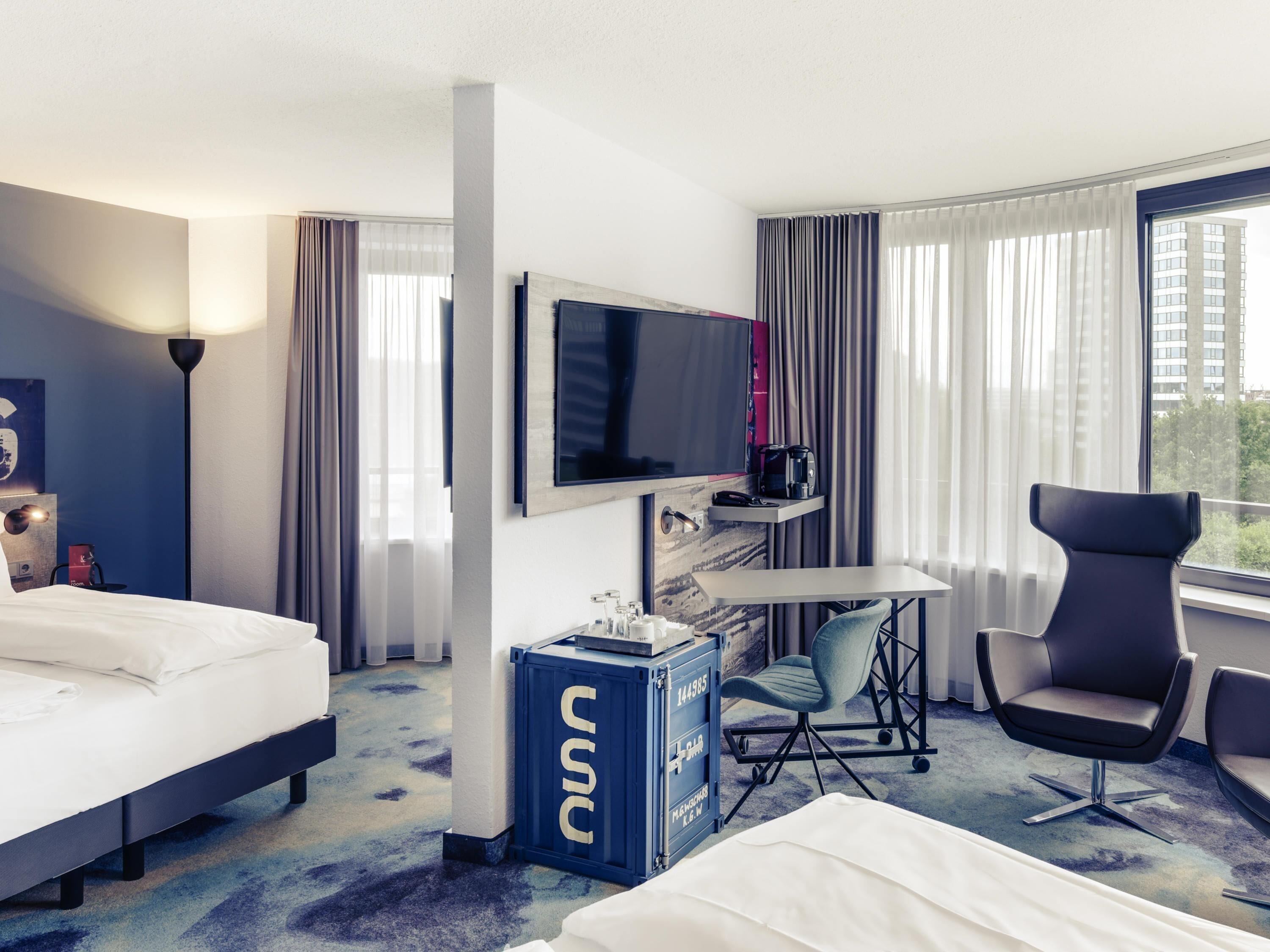 Une chambre d’hôtel avec deux lits, une télévision, un bureau avec une chaise et une grande fenêtre avec des rideaux.