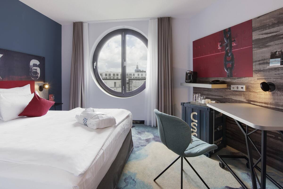 Ein Hotelzimmer mit einem Bett, einem Schreibtisch, einem Sessel und einem großen Fenster mit Blick auf Gebäude.