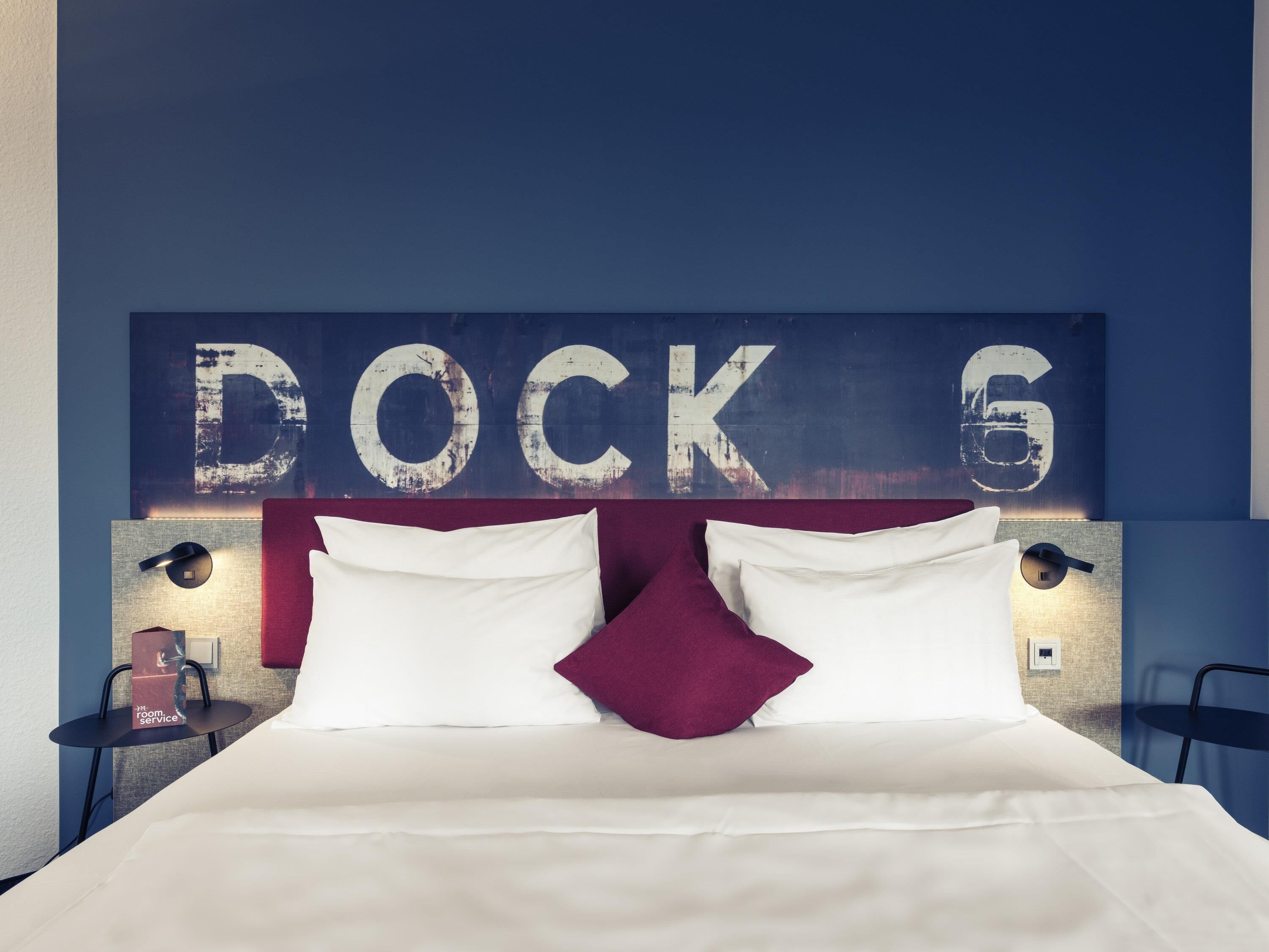 Een hotelkamer met een bed, kussens, een nachtkastje, een lamp en een muur met de tekst 'DOCK 6'.