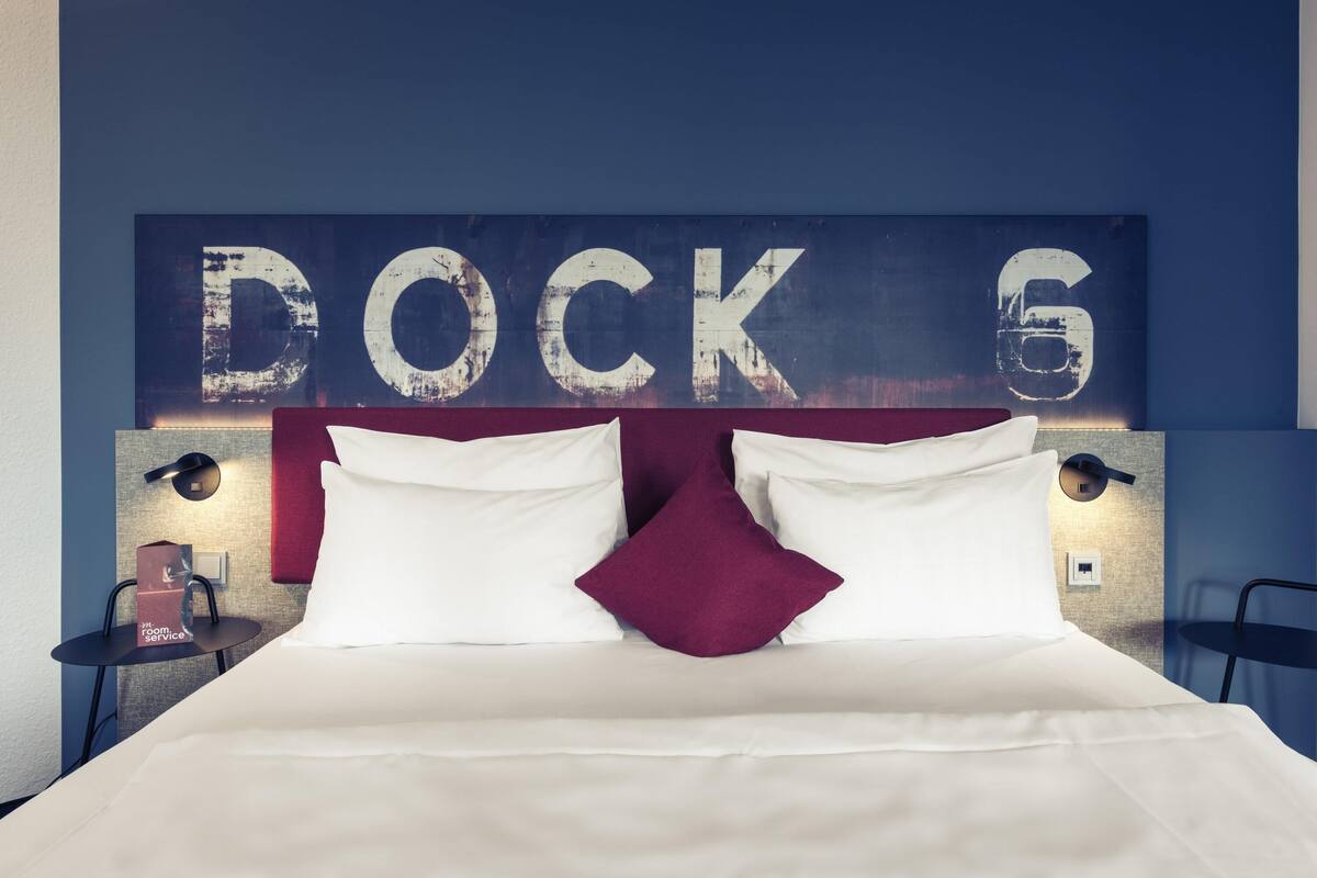 Ein Hotelzimmer mit Bett, Kissen, Nachttisch, Lampe und einer Wand mit dem Schriftzug 'DOCK 6'.