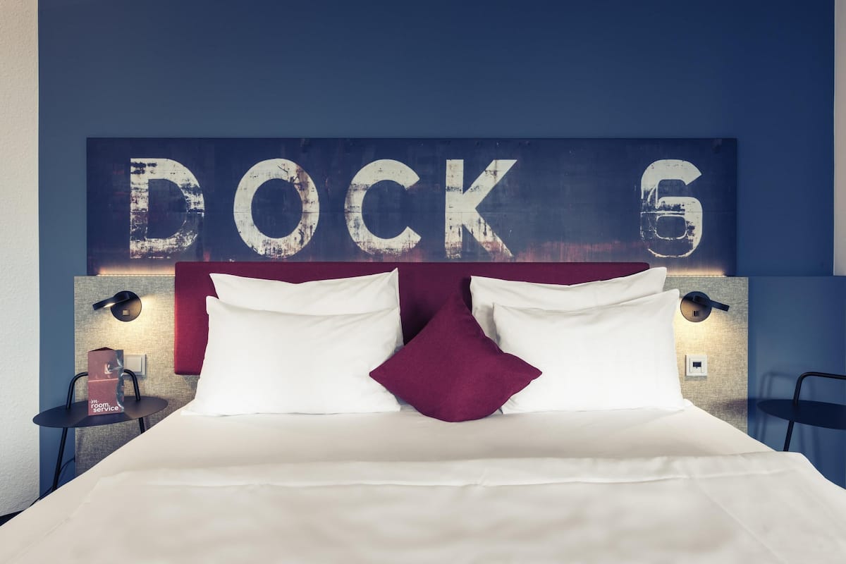A hotel room with a bed, pillows, a nightstand, a lamp, and a wall with the text 'DOCK 6'.