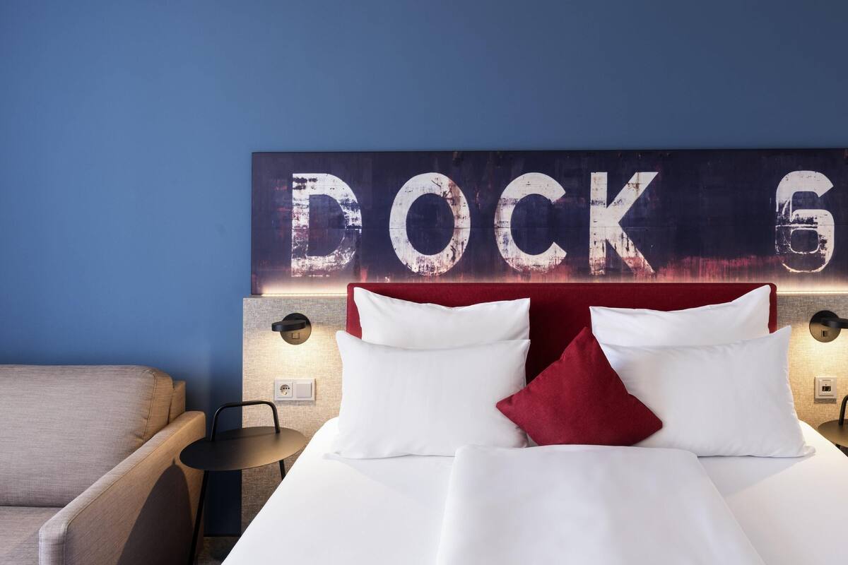Ein Hotelzimmer mit einem Bett, zwei Kissen, einem Nachttisch, einem Sessel und einer Wand mit dem Schriftzug 'DOCK 6'.