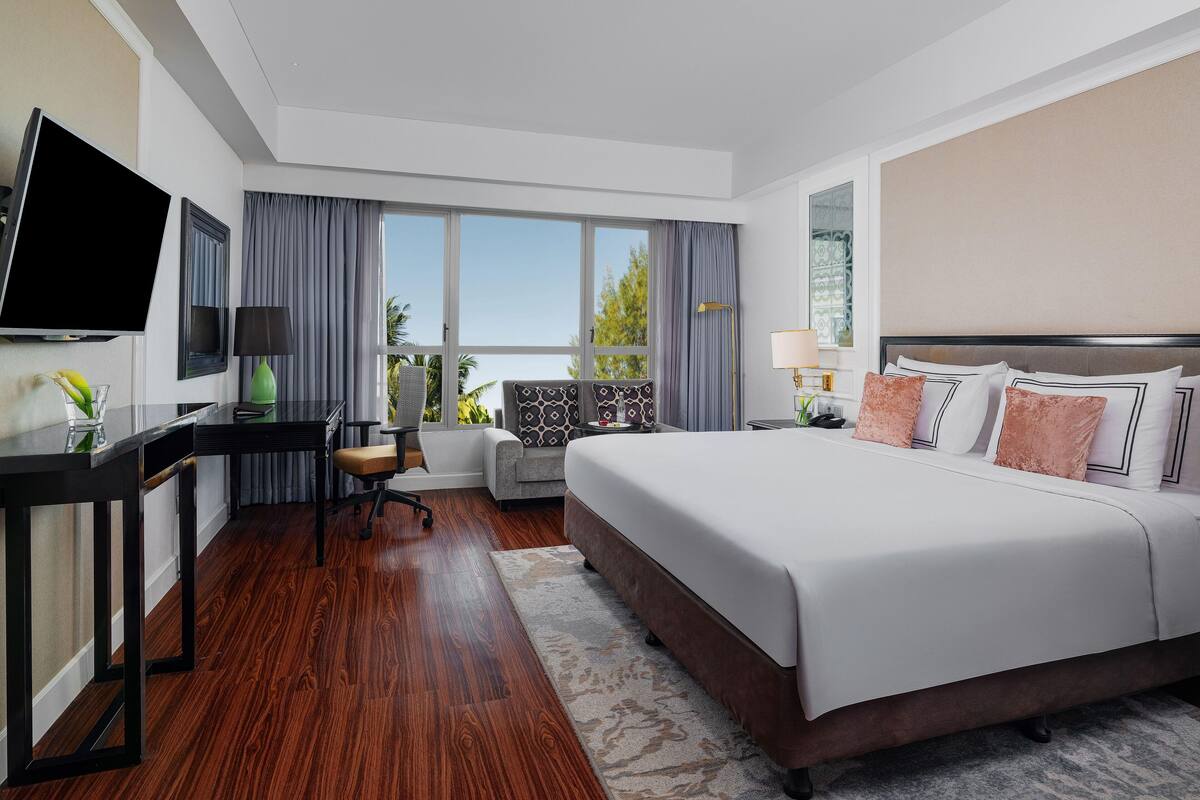 A hotel room with a large bed, a desk with a TV, a chair, a window with curtains, and a view of the outdoors.