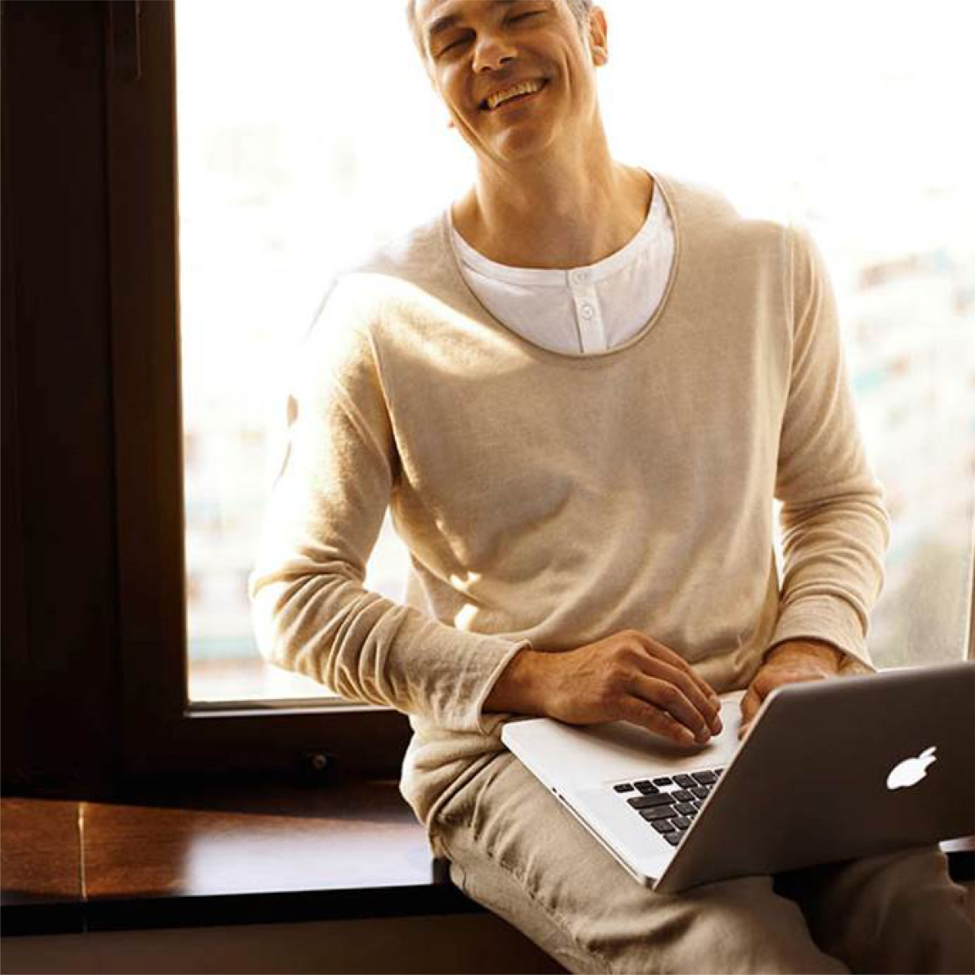Eine Person sitzt am Fenster mit einem Laptop, trägt ein weißes Langarmshirt und helle Hosen.