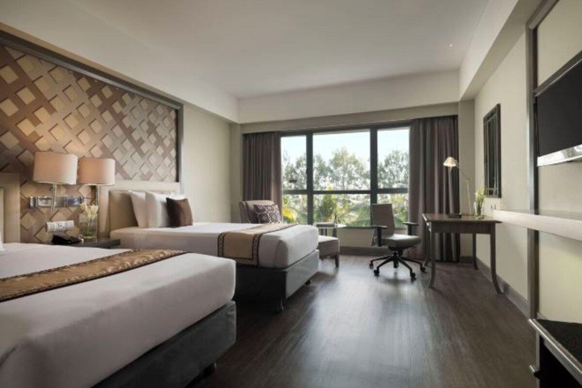 Habitación de hotel con dos camas, un escritorio, una silla, un ventanal grande y un panel decorativo en la pared.