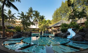 Une piscine avec un toboggan, entourée de plantes tropicales et bordée d’une architecture traditionnelle.