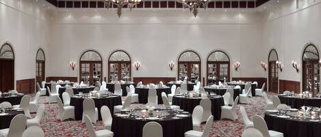 A banquet hall with multiple round tables, white chairs, chandeliers, and arched windows.