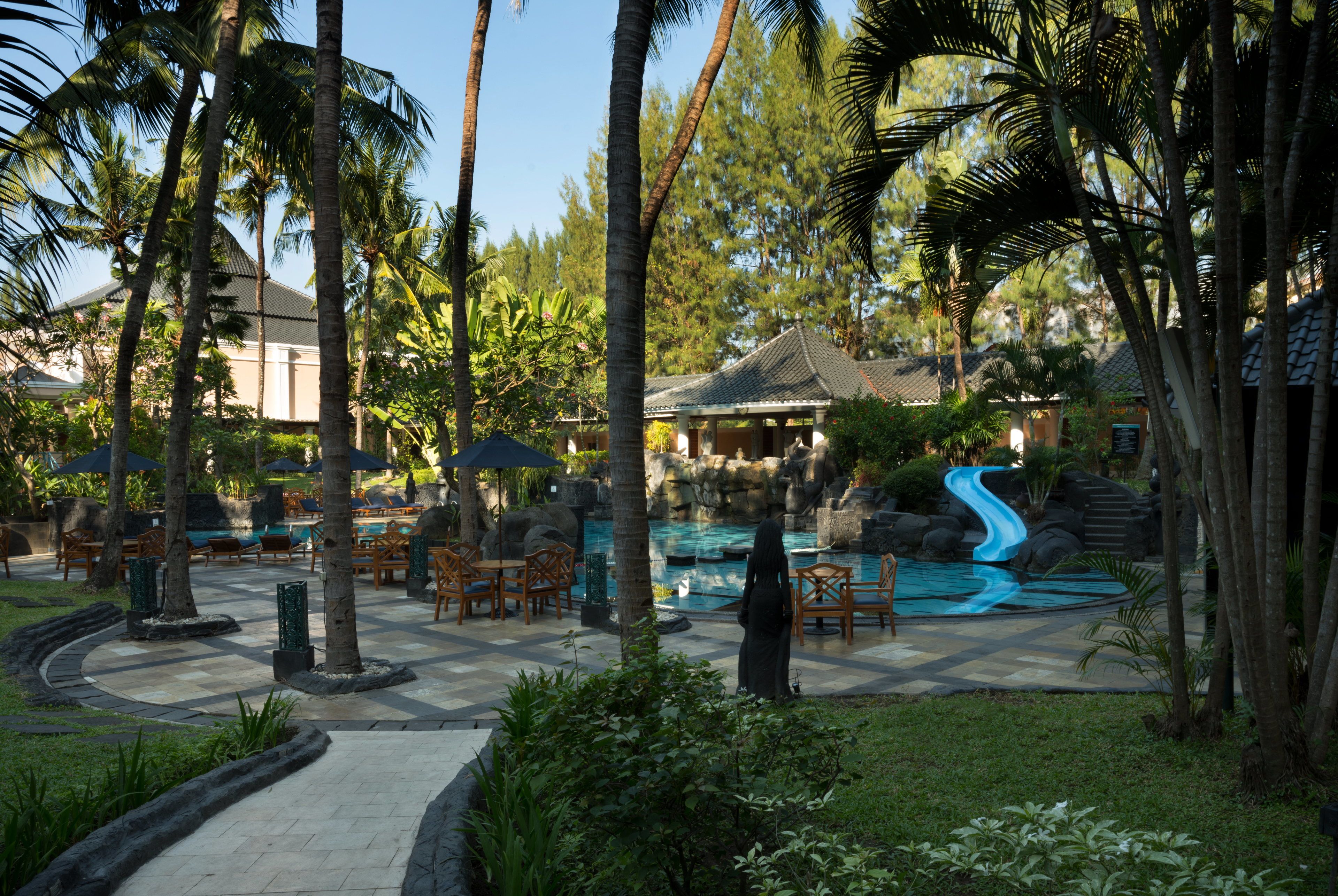 Zona de piscina en un resort con tobogán acuático, palmeras y mobiliario exterior.