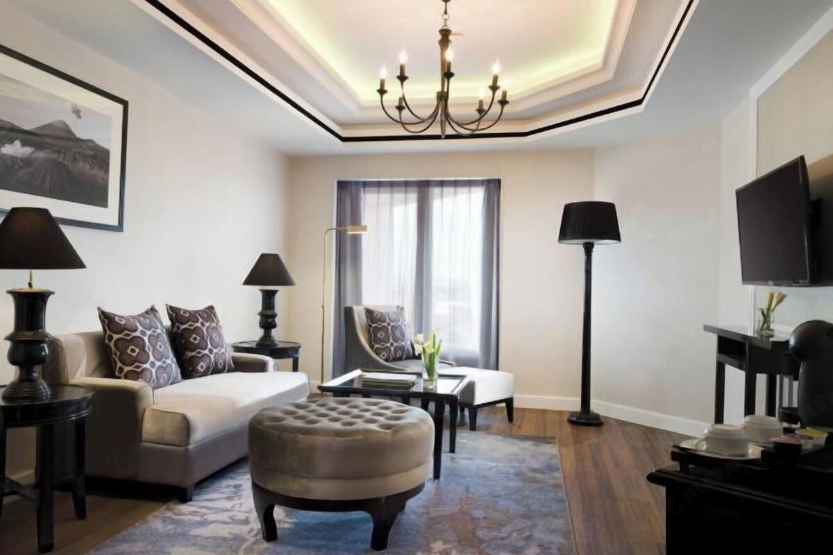 A modern hotel room with a sofa, ottoman, a flat-screen TV, a chandelier, and a large window with curtains.