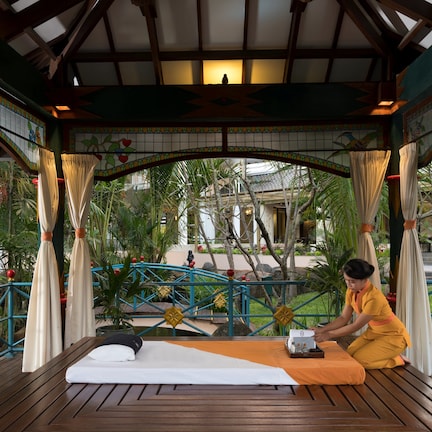 A wooden deck with a massage table, a person in a yellow robe, and a tropical garden view.