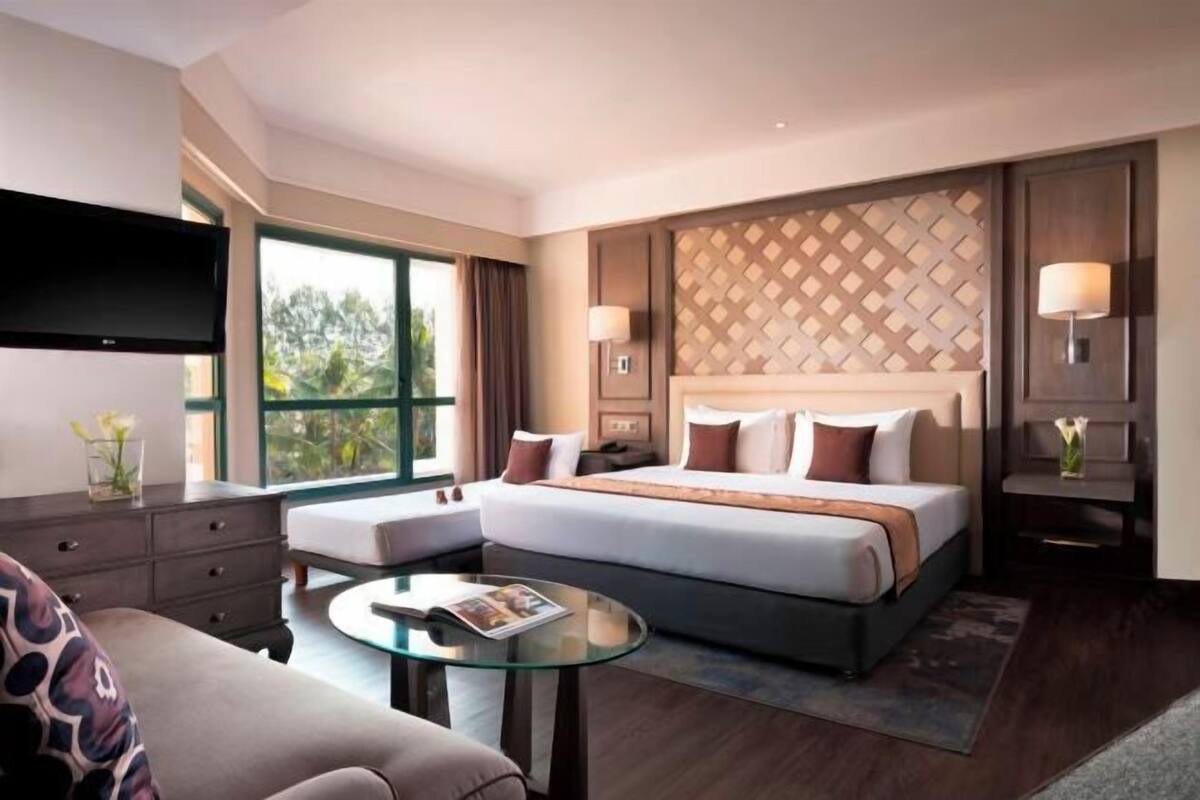 A hotel room with a bed, a TV, a sofa, a glass coffee table, and a view of the outdoors.