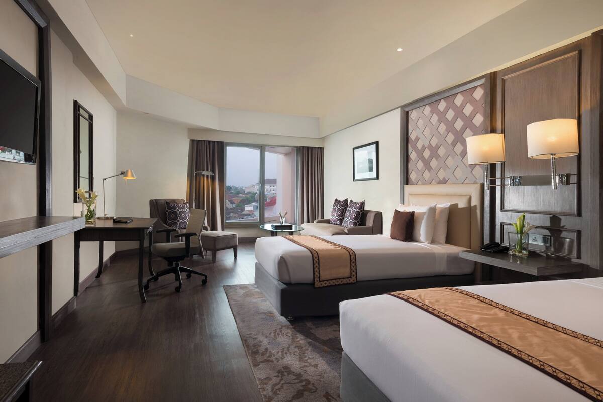 A hotel room with a large bed, a smaller bed, a desk with a chair, a sofa, and a view of the city.