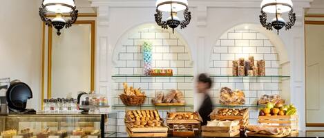 Mostrador de panadería con pasteles y pan, bajo tres candelabros, con encimera de mármol y pared de azulejos con arcos.