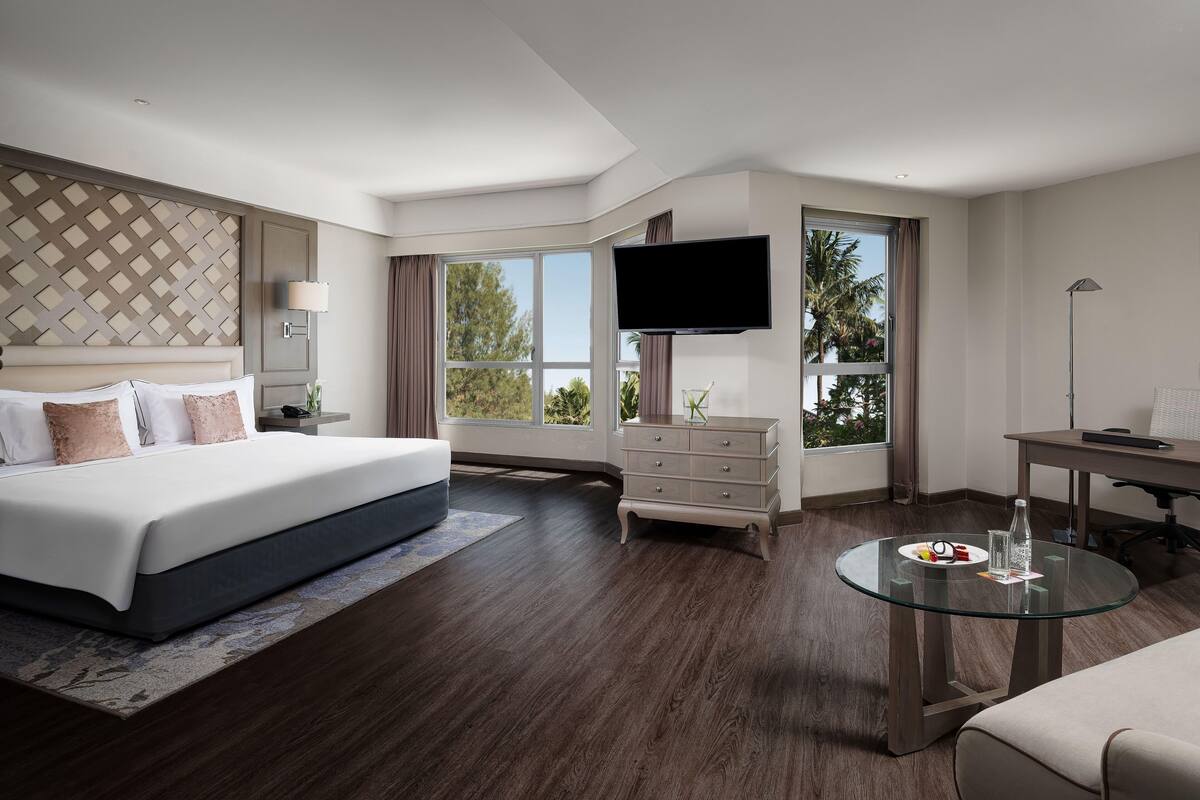 A modern hotel room with a large bed, a TV, a desk, and a glass table.