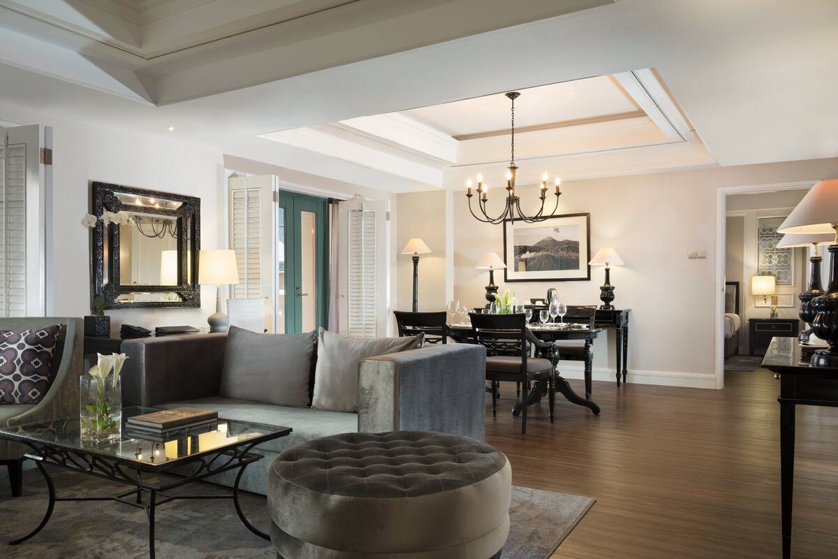 A spacious living room with a high ceiling, a dining area with a chandelier, and a cozy seating area with a tufted ottoman.