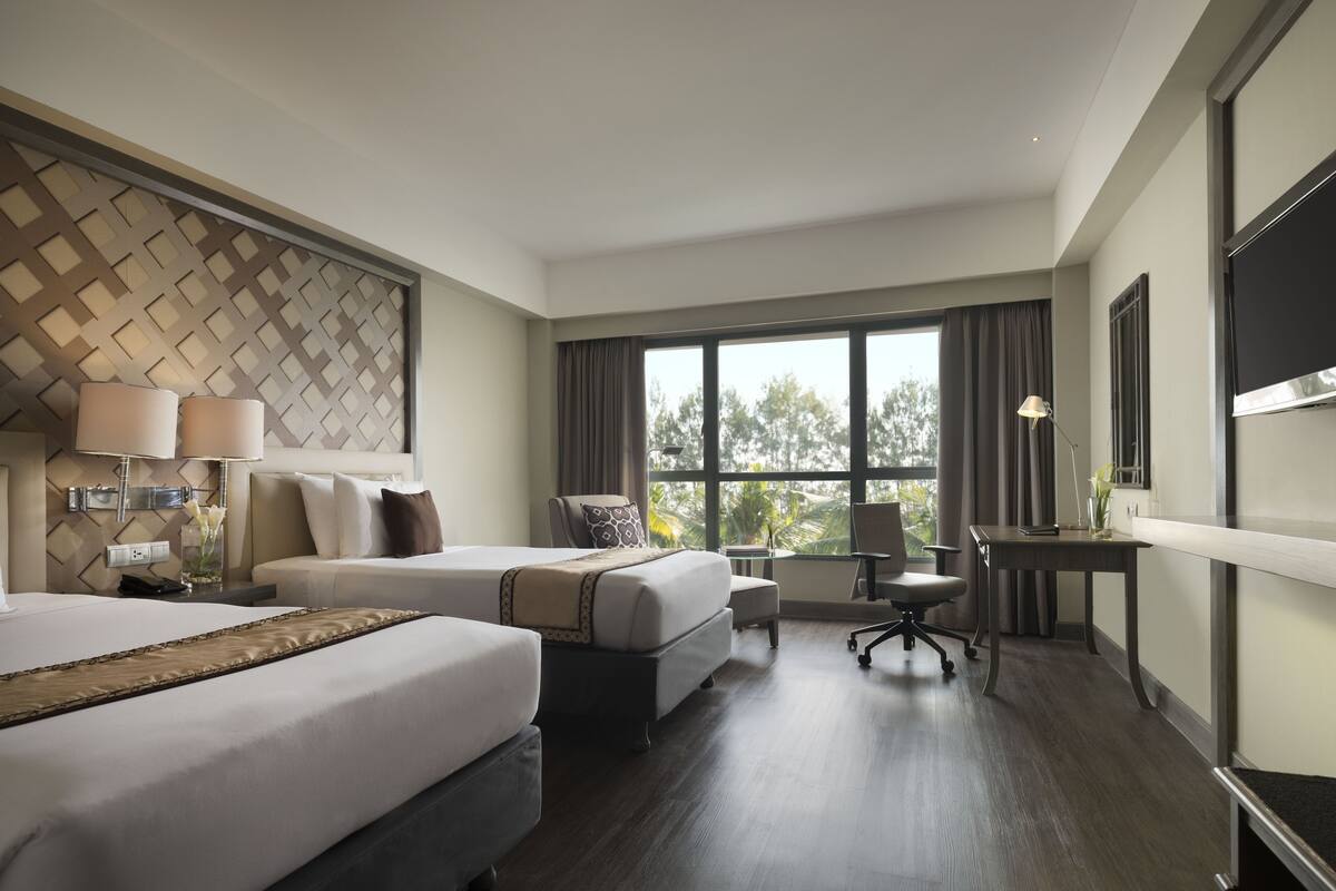 A hotel room with two beds, a desk, a chair, and a large window offering a view of trees.
