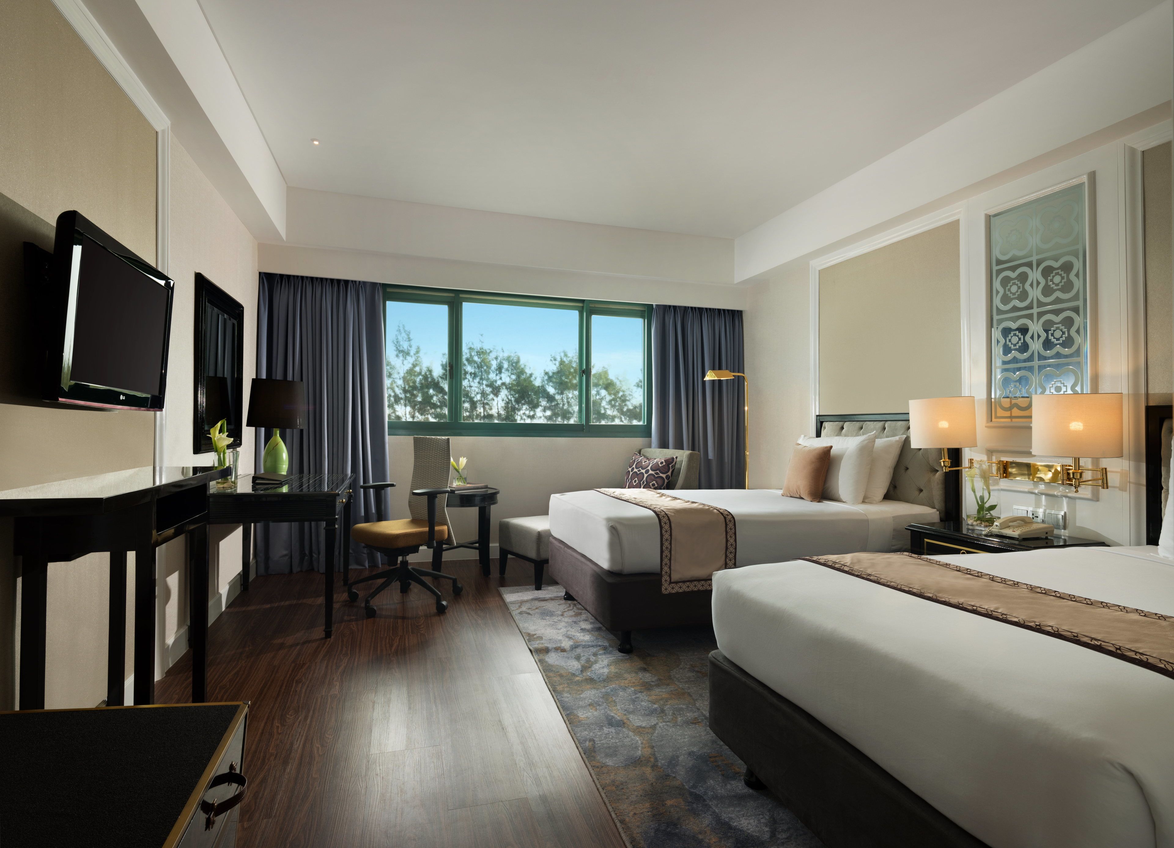A hotel room with a bed, a desk with a computer, a chair, a small table with a lamp, and a window with curtains.