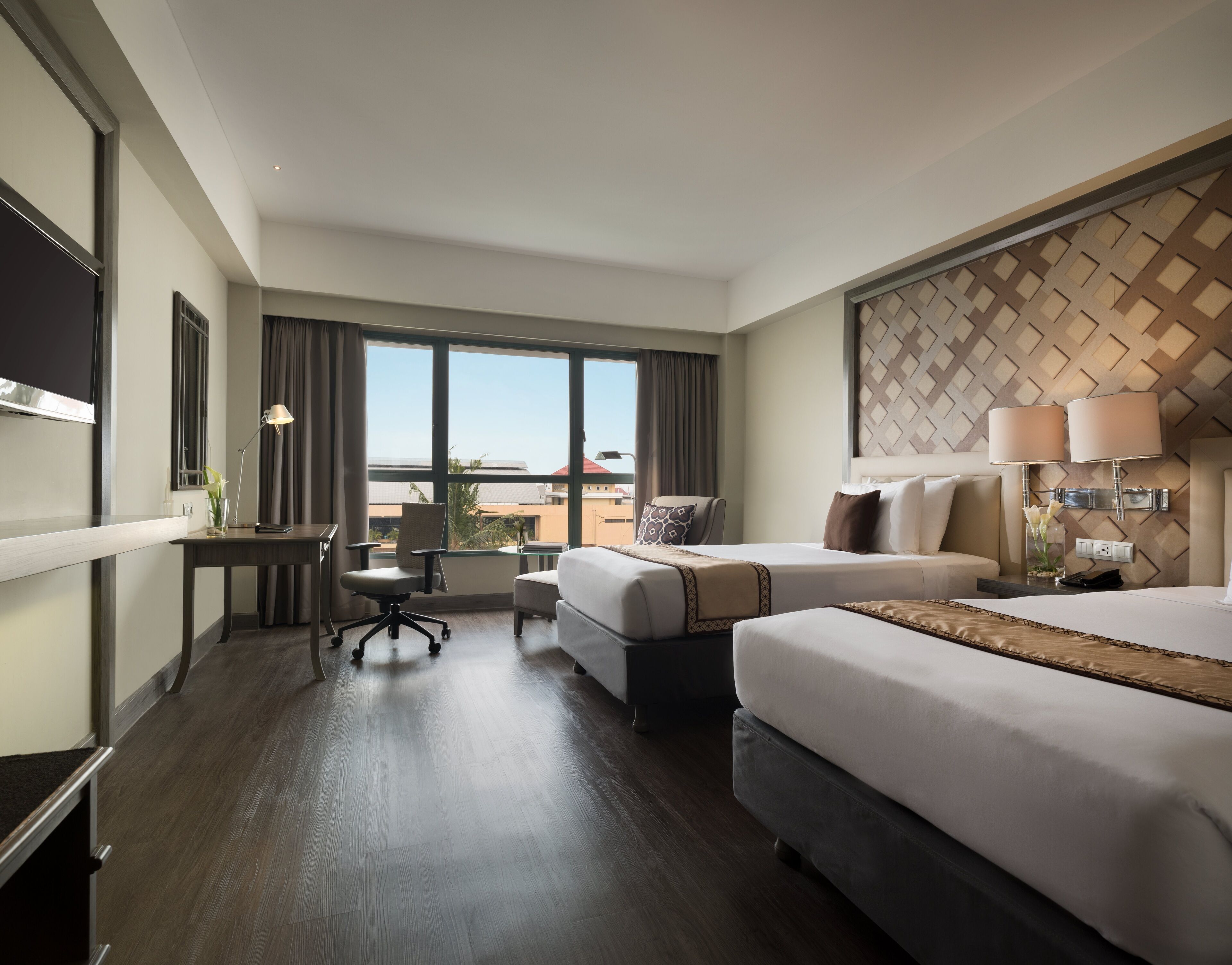 A hotel room with two beds, a desk, a chair, and a large window offering a city view.