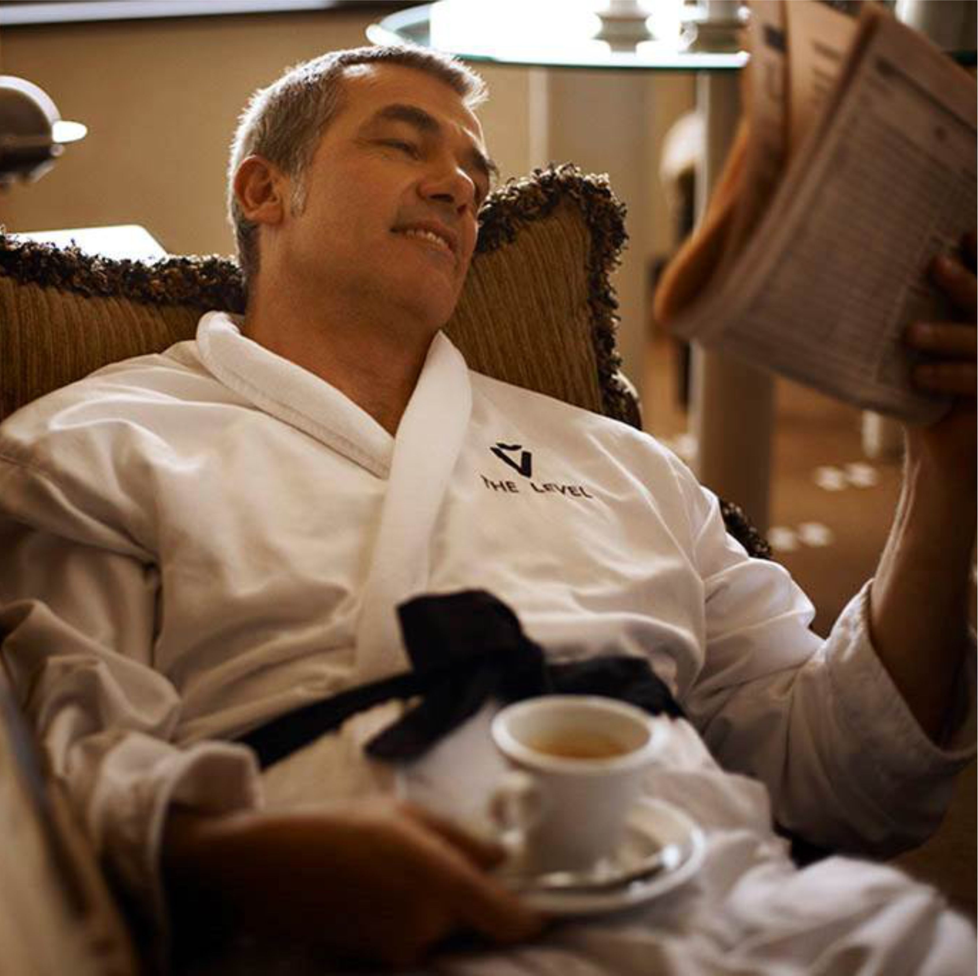 Uma pessoa vestida com um robe branco sentada em uma cadeira acolchoada, segurando uma xícara de café e um livro.