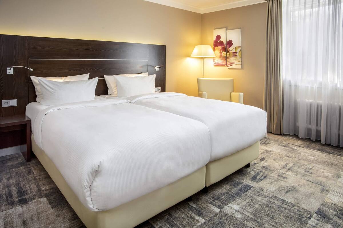A double bed with white linens, a wooden headboard, a bedside table, a lamp, and a painting on the wall.