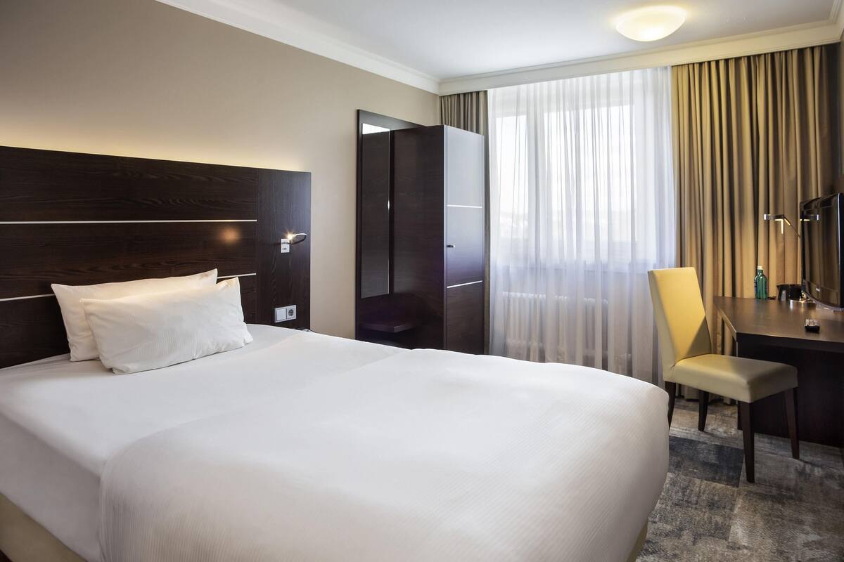 A hotel room with a large bed, a desk, a chair, a mirror, and a window with curtains.