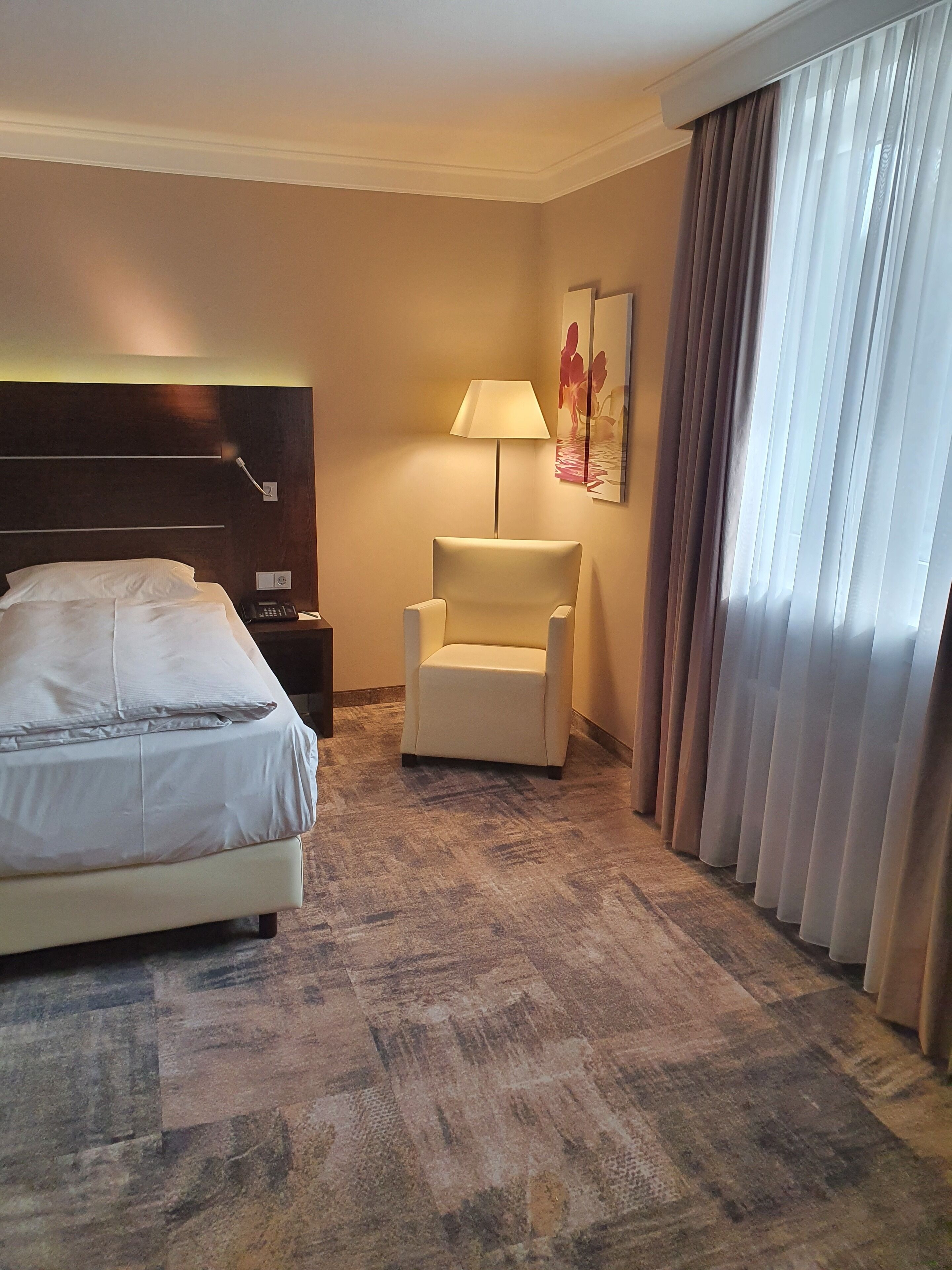 A hotel room with a bed, a chair, a lamp, and a painting on the wall.