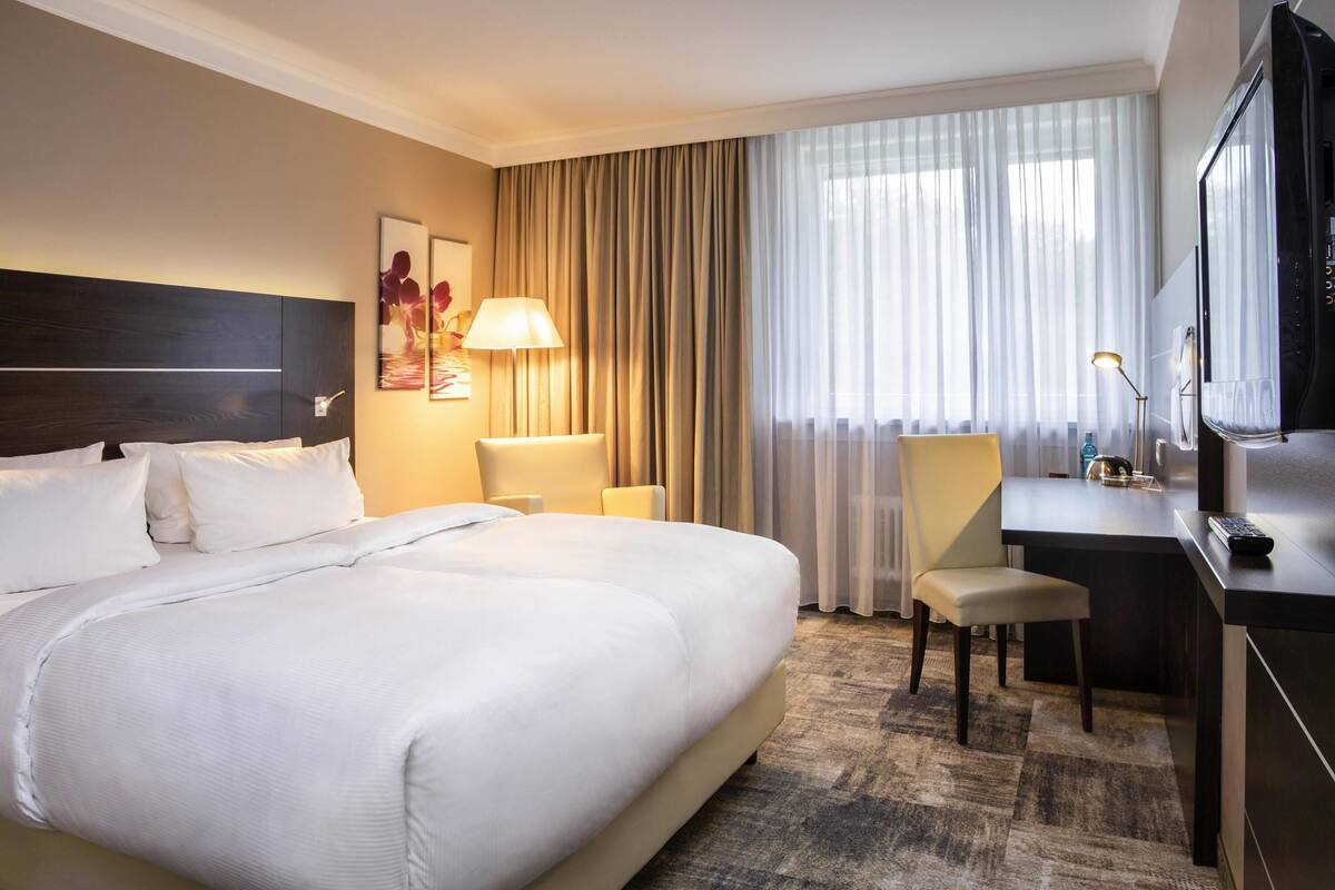 A hotel room with a large bed, a desk, a chair, a TV, and a window with curtains.