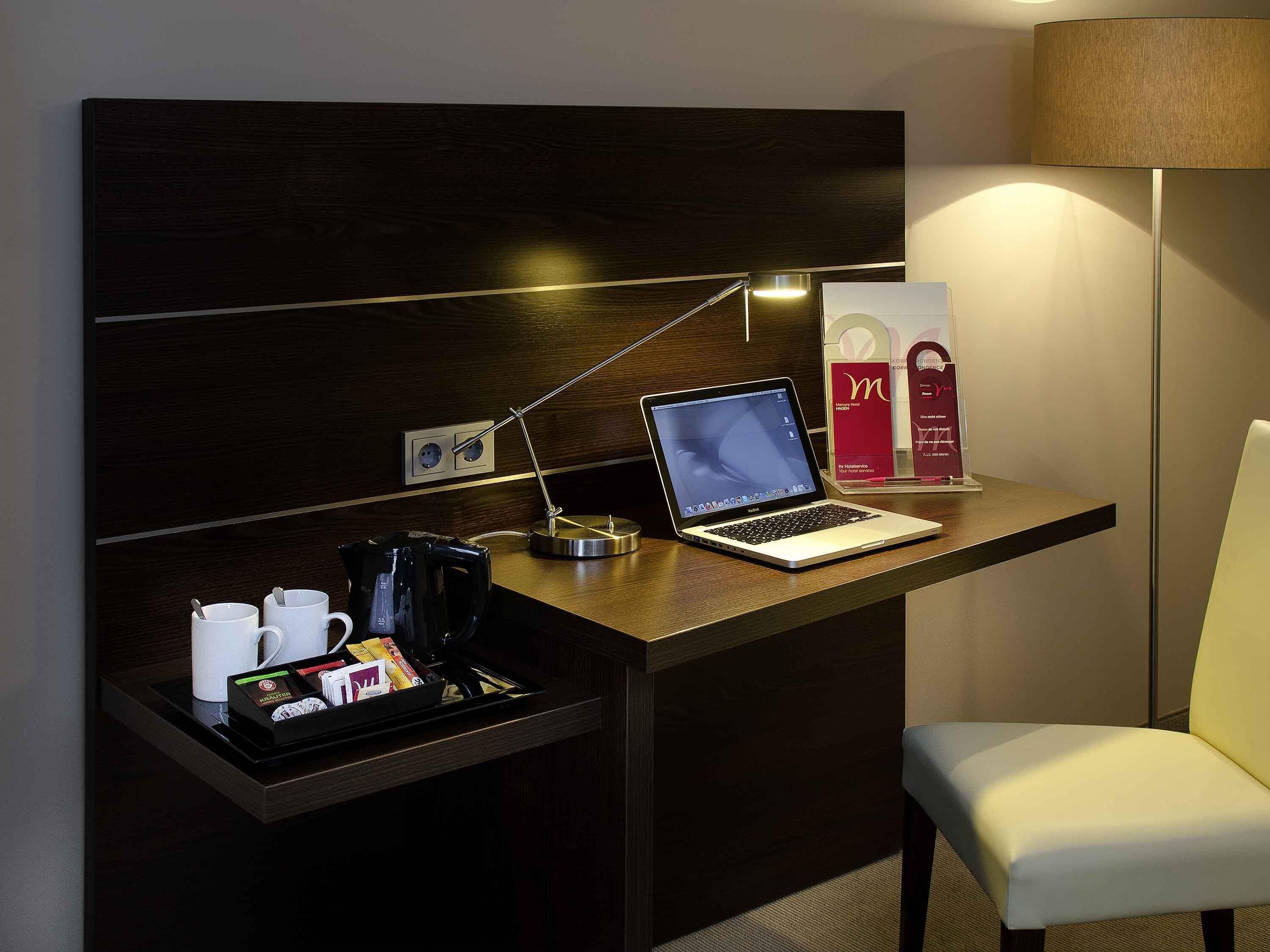 A hotel room with a desk, a laptop, a coffee maker, a tray with coffee cups and a coffee pod, a lamp, and a chair.