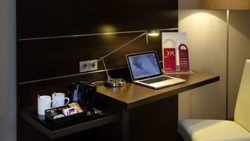 A hotel room with a desk, a laptop, a coffee maker, a tray with coffee cups and a coffee pod, a lamp, and a chair.