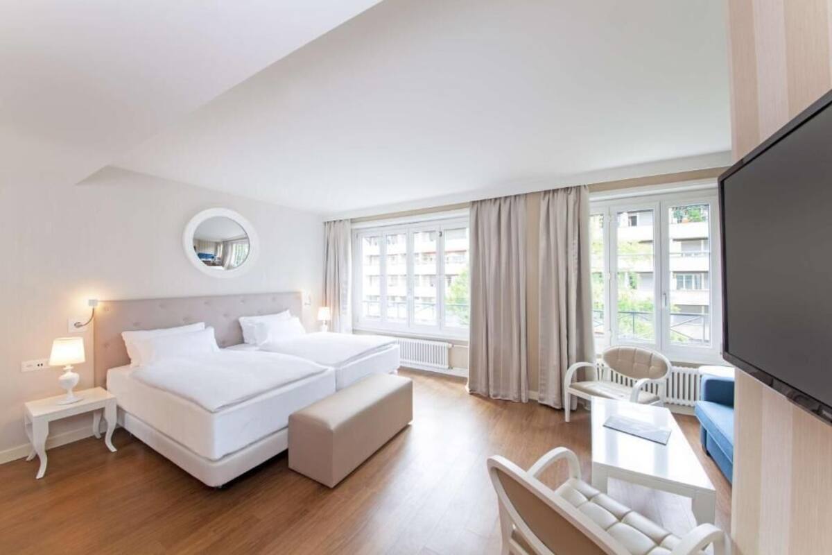 Habitación de hotel con cama doble, una mesita de noche pequeña, un espejo redondo, televisión y vistas al exterior a través de ventanas amplias.