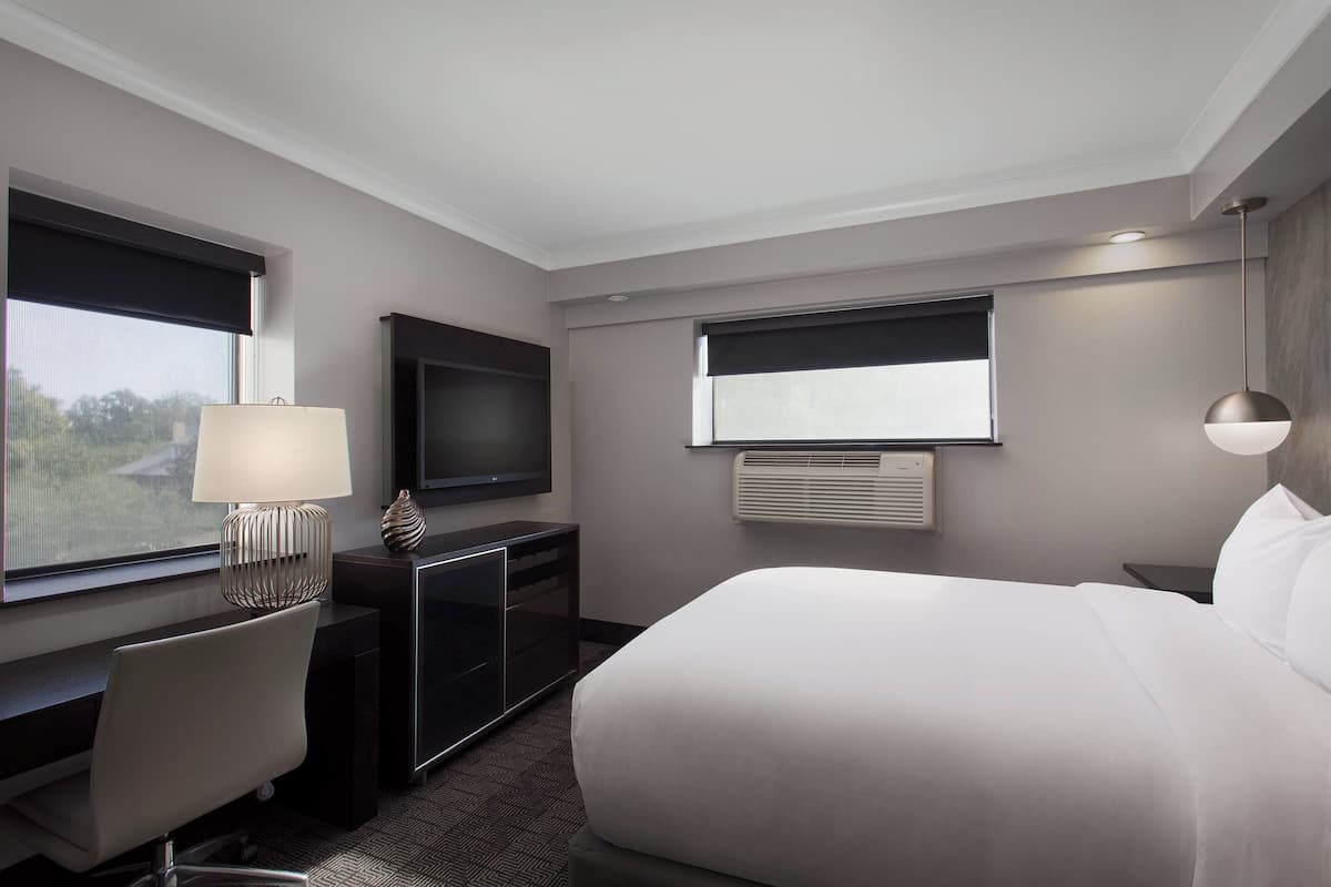 Una habitación de hotel moderna con una cama grande, un escritorio con lámpara, un televisor de pantalla plana y un ventanal con persianas.