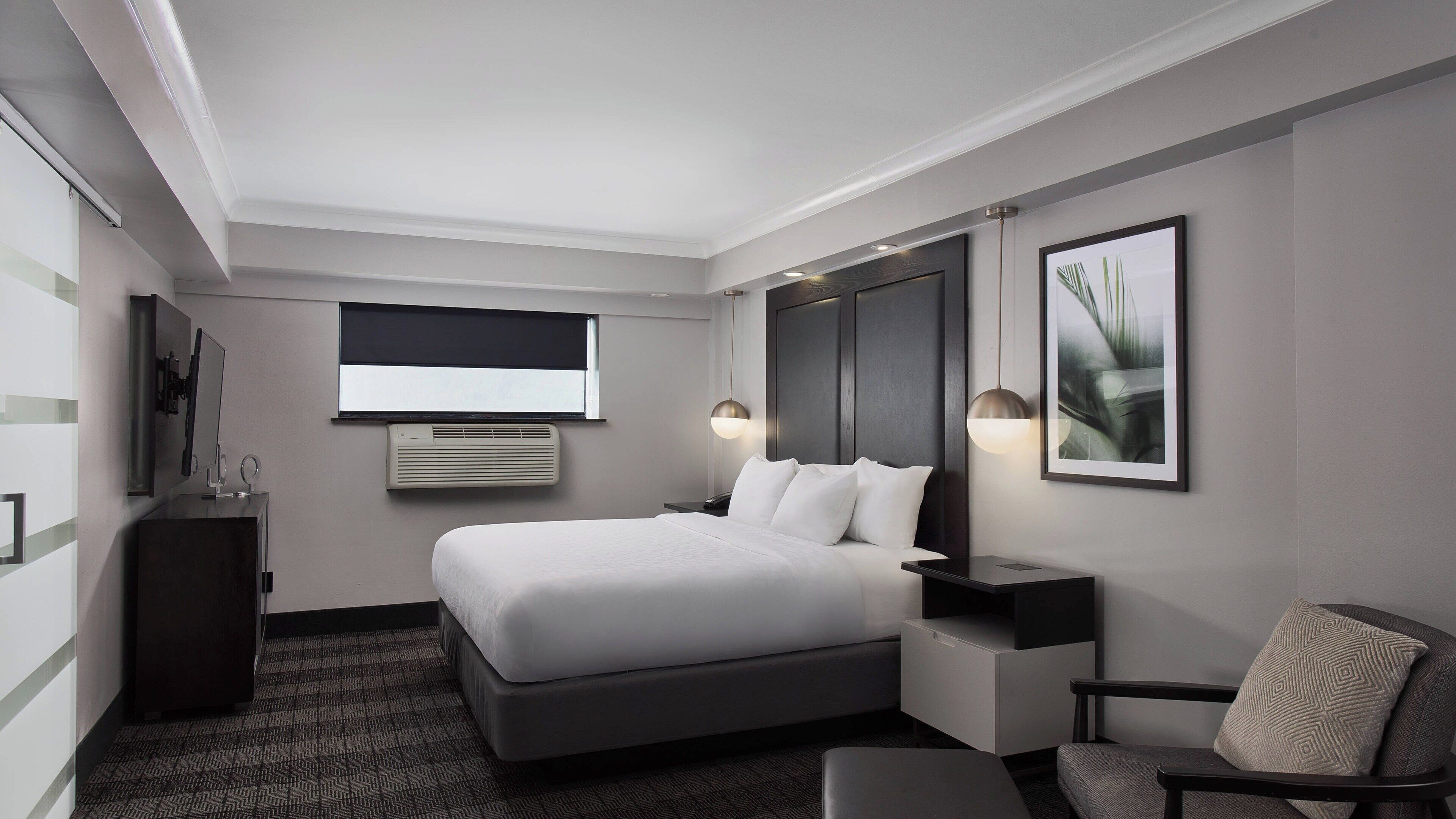 A modern hotel room with a large bed, a desk, a chair, a TV, and a window with blinds.
