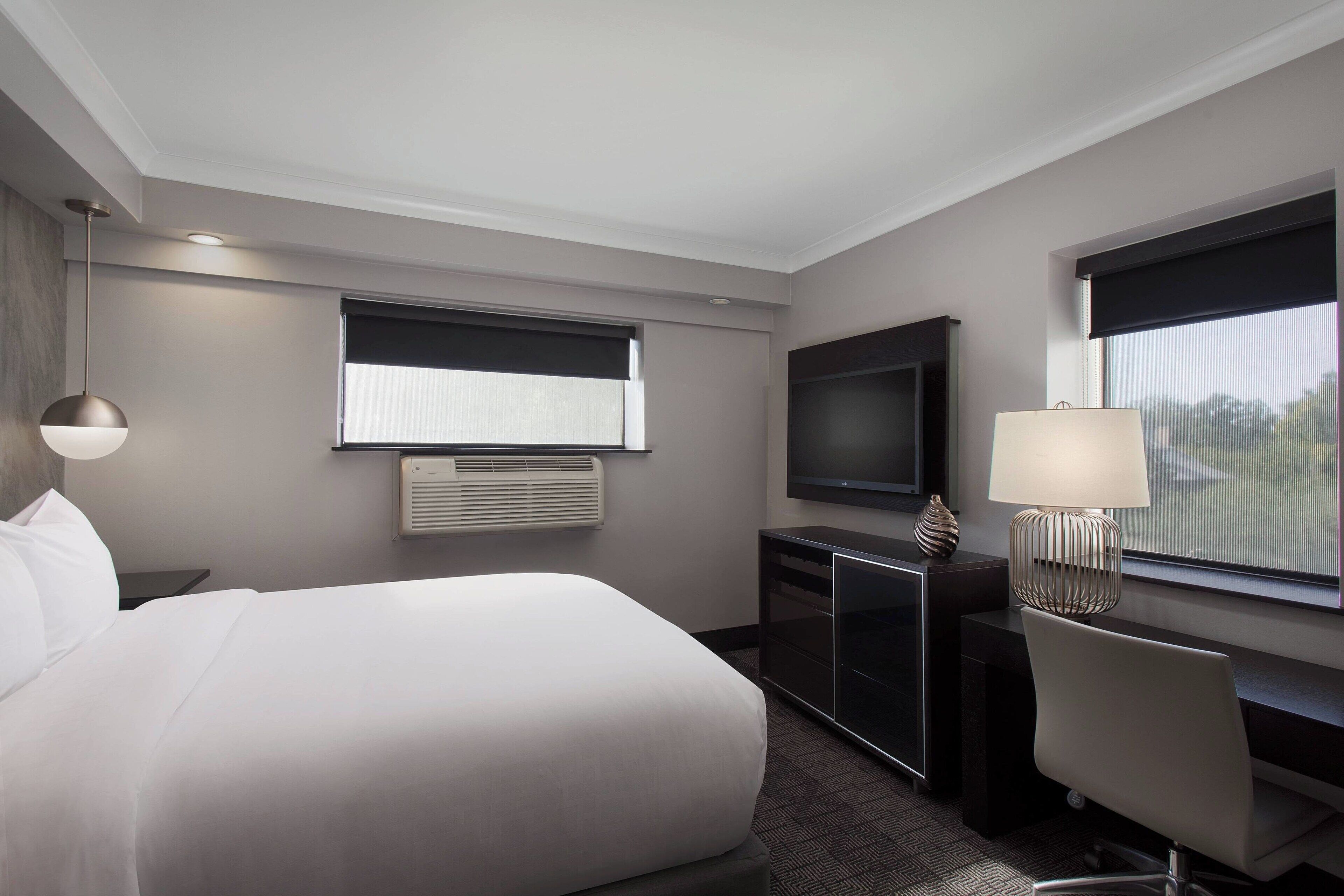 A modern hotel room with a large bed, a desk, a chair, a TV, and a window with blinds.