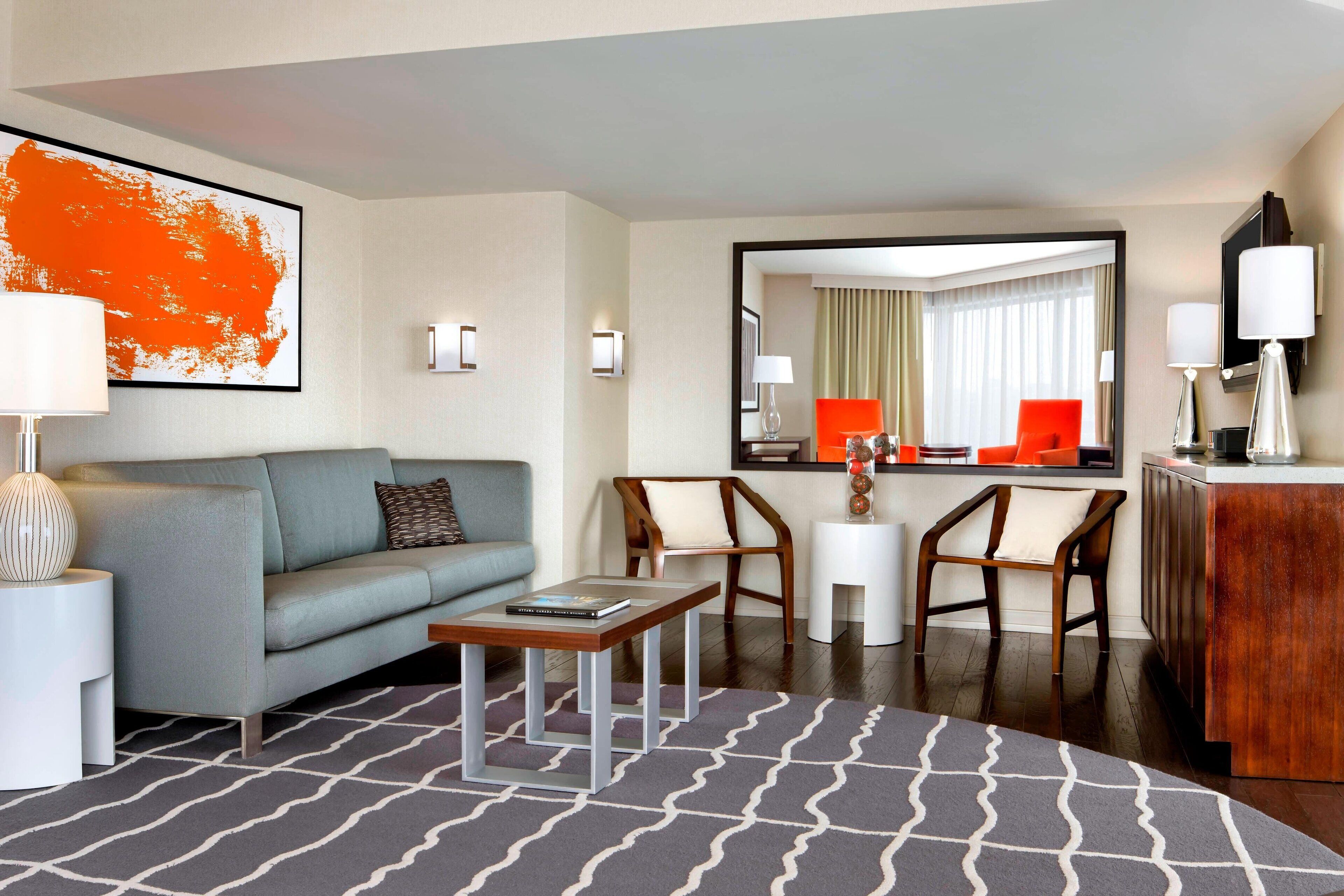 A modern hotel room with a grey sofa, a wooden coffee table, two wooden chairs, a small white side table, a large mirror, and a wooden cabinet.