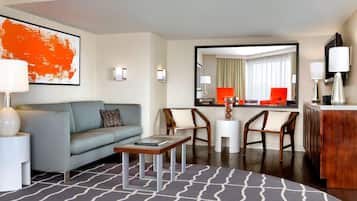 A modern hotel room with a grey sofa, a wooden coffee table, two wooden chairs, a small white side table, a large mirror, and a wooden cabinet.