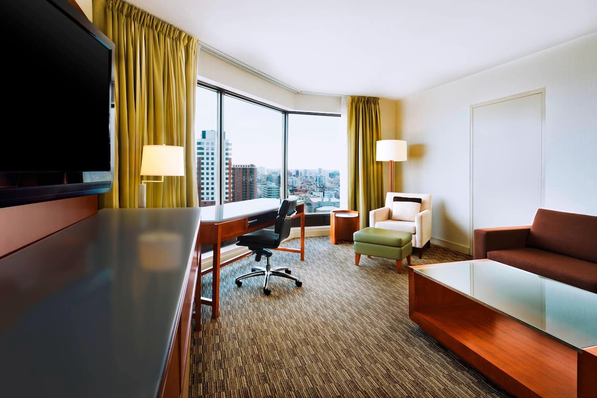 A hotel room with a large window, a flat-screen TV, a desk with a chair, a sofa, and a lamp.