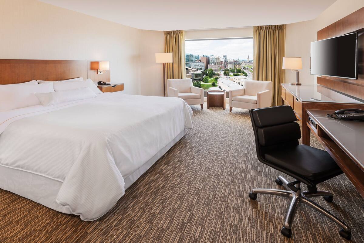 A hotel room with a large bed, a desk with a chair, a TV, and a view of the city.
