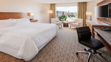 A hotel room with a large bed, a desk with a chair, a TV, and a view of the city.