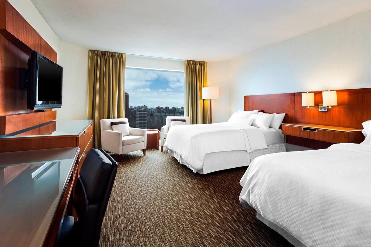 A hotel room with a large bed, a smaller bed, a desk with a TV, a chair, a lamp, and a view of the city.