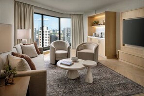 Habitación de hotel moderna con sofá, dos sillones, una mesita de centro, televisor de pantalla plana grande, zona de cocina y vistas a la ciudad a través de amplias ventanas.